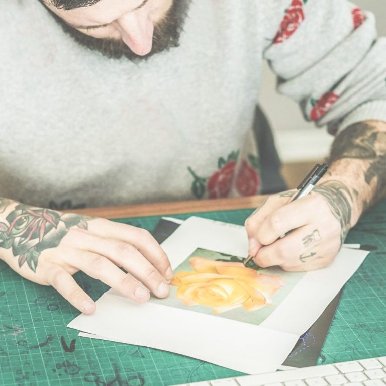 Eine männliche tätowierte Person zeichnet eine orange Rose auf ein weißes Blatt Papier