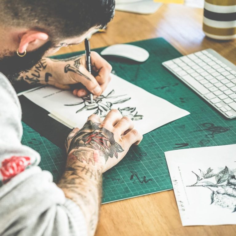 Ein tätowierter Mann fertigt eine Tattoo Vorlage auf einem Schreibtisch an.